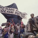 A foto mostra a escultura "Cristo Mendigo" que foi o carro abre-alas do desfile Ratos e Urubus. Na imagem, a escultura é coberta por sacos de lixo como uma censura, e carrega a faixa que diz "Mesmo proibido olhai por nós!"