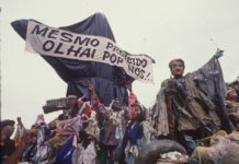 A foto mostra a escultura "Cristo Mendigo" que foi o carro abre-alas do desfile Ratos e Urubus. Na imagem, a escultura é coberta por sacos de lixo como uma censura, e carrega a faixa que diz "Mesmo proibido olhai por nós!"