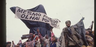 A foto mostra a escultura "Cristo Mendigo" que foi o carro abre-alas do desfile Ratos e Urubus. Na imagem, a escultura é coberta por sacos de lixo como uma censura, e carrega a faixa que diz "Mesmo proibido olhai por nós!"