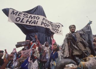 A foto mostra a escultura "Cristo Mendigo" que foi o carro abre-alas do desfile Ratos e Urubus. Na imagem, a escultura é coberta por sacos de lixo como uma censura, e carrega a faixa que diz "Mesmo proibido olhai por nós!"