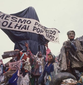 A foto mostra a escultura "Cristo Mendigo" que foi o carro abre-alas do desfile Ratos e Urubus. Na imagem, a escultura é coberta por sacos de lixo como uma censura, e carrega a faixa que diz "Mesmo proibido olhai por nós!"