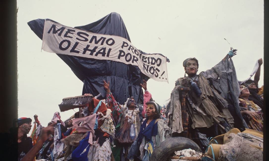 A foto mostra a escultura "Cristo Mendigo" que foi o carro abre-alas do desfile Ratos e Urubus. Na imagem, a escultura é coberta por sacos de lixo como uma censura, e carrega a faixa que diz "Mesmo proibido olhai por nós!"