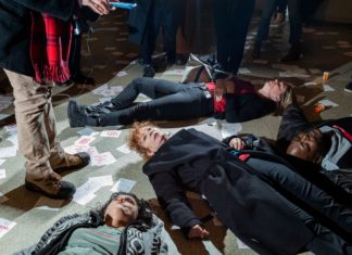 A foto mostra protestantes performance um "Die in". A manifestação é contra a família Sackler, ligado à Purdue Pharma, produtora do Oxy Contin. Em meio aos protestantes está a fotógrafa Nan Goldin, rodeada por papéis que dizem "Sackler Lie People Die" e frascos de remédio