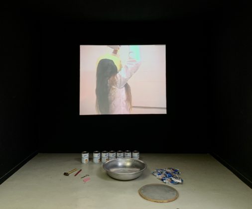 Peça da exposição Ratos e Urubus - A foto mostra a exibição de uma gravação da performance "Pancake" de Márcia X. Abaixo do telão onde é projetado o vídeo estão objetos equivalentes aos usados por ela na sua performance.