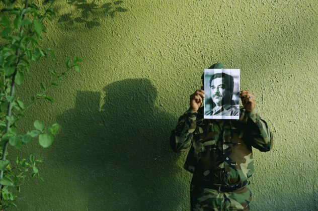 Na foto, trabalho da série "Saddam is Here" de Jamal Peniweny, vemos um homem, posicionado contra uma parede verde, vestido com roupas militares que segura uma foto impressa em papel comum de Saddam Hussein