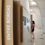 No primeiro plano, divisões de madeira na parede tem o escrito "Entre Bordas", na vertical. Ao fundo, uma mulher olha quadros da exposição "Entre bordas - sons que escapam" na parede