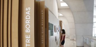 No primeiro plano, divisões de madeira na parede tem o escrito "Entre Bordas", na vertical. Ao fundo, uma mulher olha quadros da exposição "Entre bordas - sons que escapam" na parede