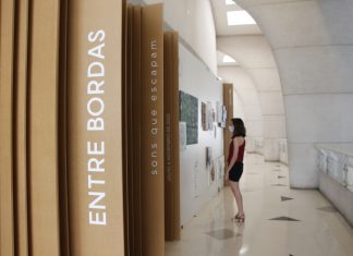 No primeiro plano, divisões de madeira na parede tem o escrito "Entre Bordas", na vertical. Ao fundo, uma mulher olha quadros da exposição "Entre bordas - sons que escapam" na parede