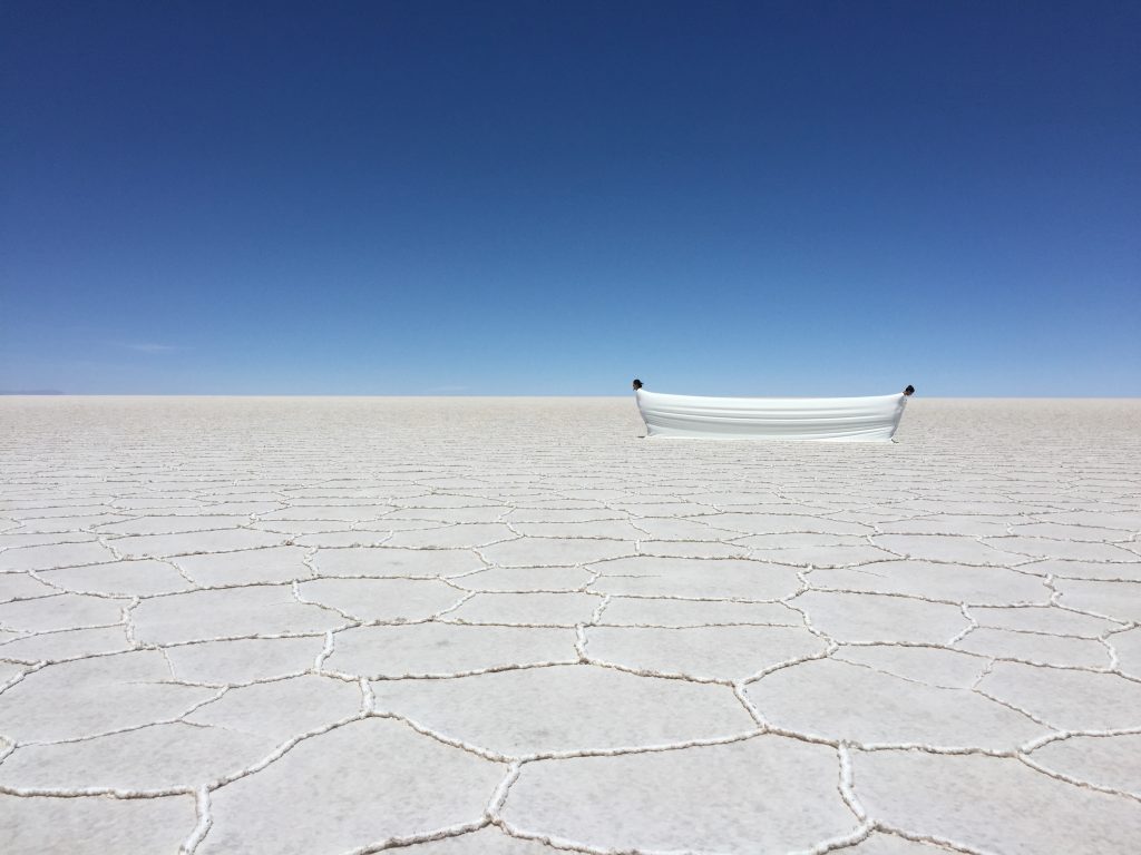 A imagem mostra a obra "Estudo para o Tempo Suspenso Ma 間 :: Silêncio, o espaço tempo de resistência e resiliência", de Dudu Tsuda, onde duas pessoas se equilibram envolvidas por um mesmo tecido, no deserto de sal 'Salar del Uyuni' para IX Biennal SIART de La Paz