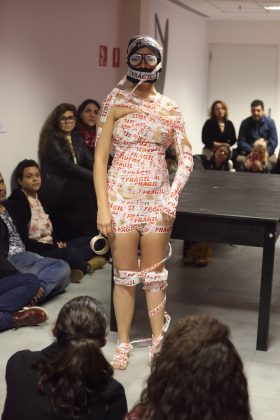 Fotografia vertical, colorida. Estela Lapponi está de pé no centro. O público, majoritariamente formado por mulheres está sentado em torno dela e a olha. Ela tem o corpo enfaixado da púbis até a cabeça com uma fita adesiva branca com escrito FRÁGIL em vermelho. Na cabeça, uma touca preta e os óculos Zuleika Brit, que possui olhos desenhados. Essa foto foi tirada pelo fotógrafo Ivson Miranda durante a performance INTENTO 00035 - ÇA M'ÉNERVE!!! do projeto Corpo Intruso apresentada no Itaú Cultural.