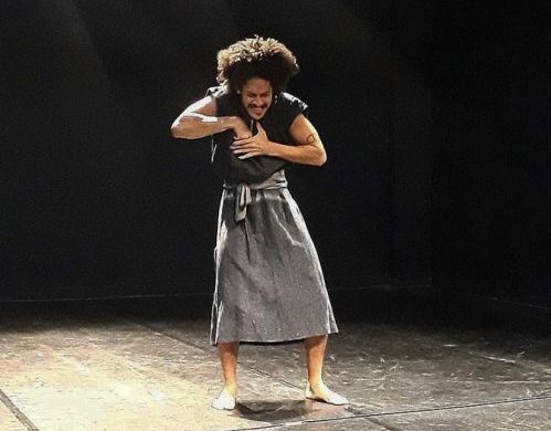 Foto horizontal, colorida. Leonardo Castilho está de pé sobre um palco sem cenário, com paredes pretas. Está iluminado por um refletor. A mão direita está sobre o peito, com os dedos apontados para baixo. A mão esquerda, aberta, também sobre o peito, cobre a direita. Tem os ombros levemente elevados e tensionados, intensificando a expressão de dor estampada em seu rosto. Ele está vestido com uma regata preta e uma saia cinza, tem os pés descalços. Fotografia de apresentação de um poema no Slam do Corpo.