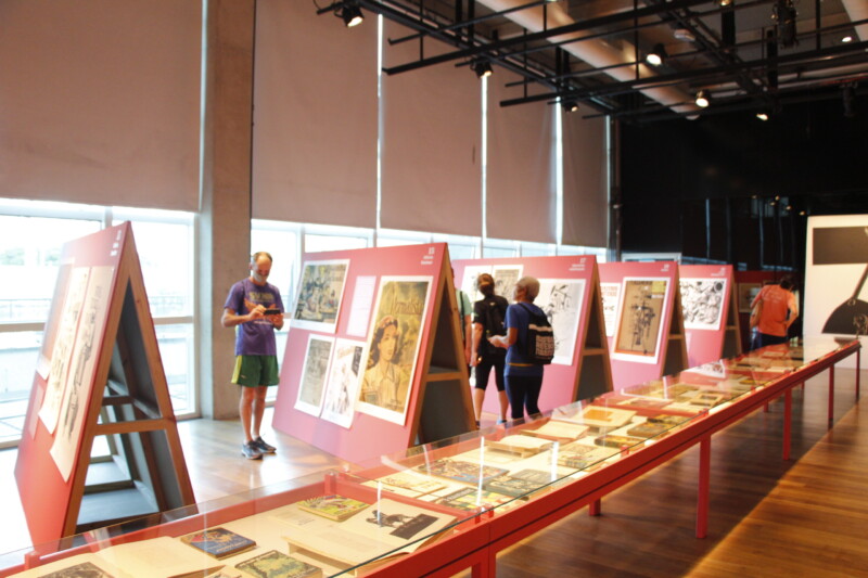 Foto horizontal, colorida. Vista da exposição TRABALHADORES ILUSTRADOS. Ao fundo, pessoas contemplam os painéis onde estão dispostas as ilustrações. À frente delas, uma mesa com os livros e revistas onde constam as ilustrações originais.