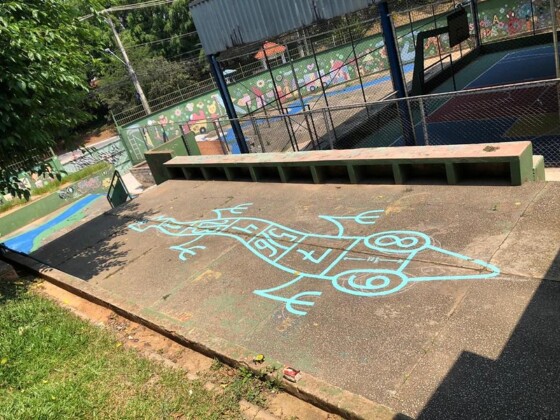 Foto horizontal, colorida. Chão de cimento de uma escola pública de São Paulo traz um trabalho do artista urbano TEC em azul, O desenho é lagarto, em tom de azul claro, formando com seu corpo atípica brincadeira amarelinha. A obra é parte do projeto Escola Criativa do Instituto Choque Cultural.