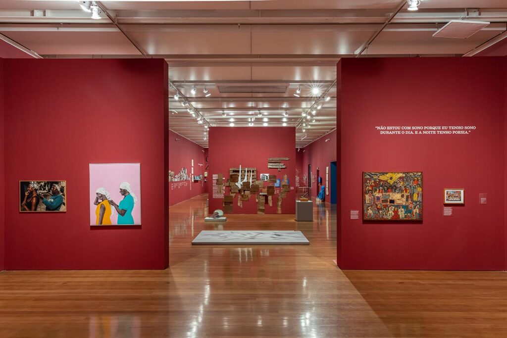 Foto horizontal, colorida. Vista da exposição CAROLINA MARIA DE JESUS: UM BRASIL PARA OS BRASILEIROS, no Instituto Moreira Salles, ilustra a cenografia da mostra a qual Fabio Cypriano se refere. As paredes vermelhas tem quadros e textos curtos, nota-se obras no chão à distância.