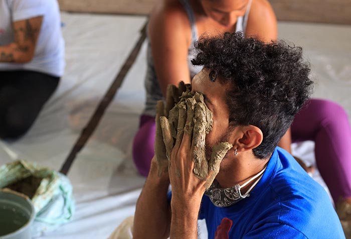 Foto horizontal, colorida. Um jovem, sentado no chão do ateliê na Residência Belojardim, leva as mãos, com argila, aos olhos. Cena é parte do projeto Corpos de Phonosophia. 