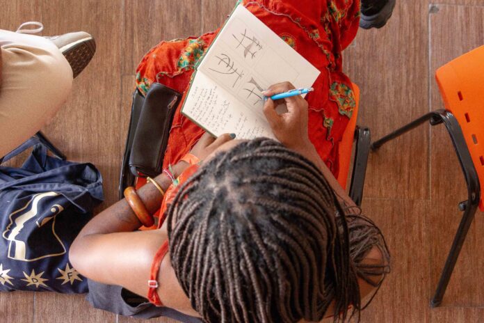 Foto horizontal, colorida. Registro da última edição de Germinadora, residência do MAM Rio voltada a estudantes de 16 a 18 anos, das redes pública e privada. Na imagem, uma adolescente sentada faz anotações e rascunhos de desenho em um caderno.